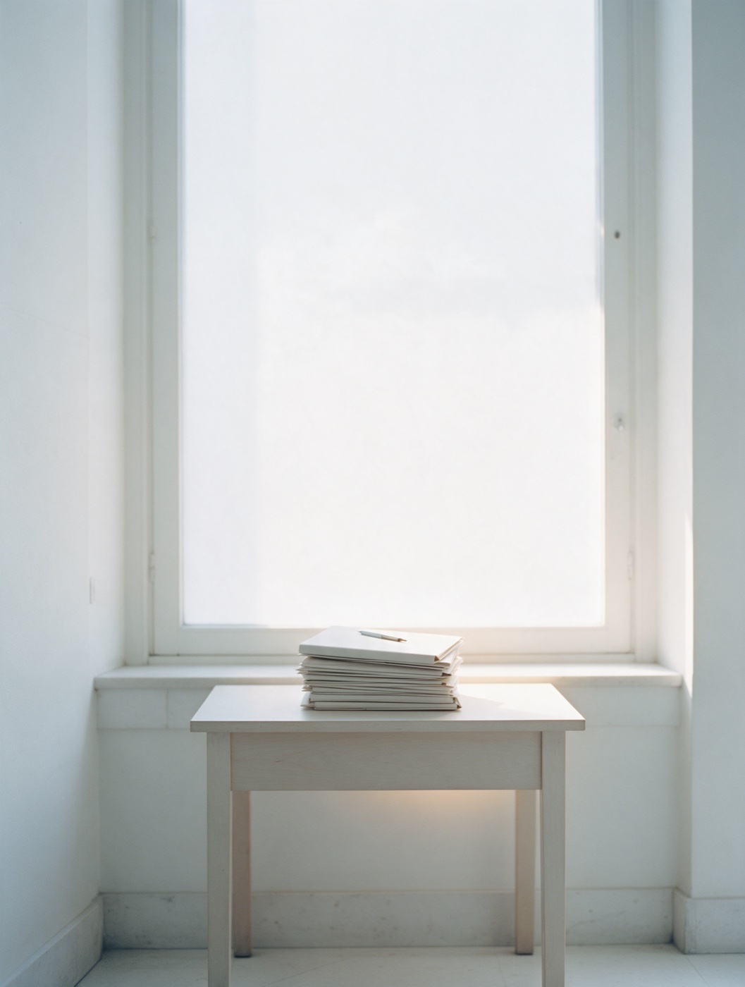 Ein einzelner Schreibtisch am Fenster, ein Stapel geschlossener Ordner, weiches Tageslicht.